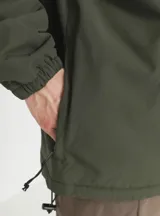 Campera tipo windbreaker de estilo pullover, color verde militar, con capucha ajustable, cierre frontal parcial y bolsillo tipo canguro con cierre. Presenta el logo de la marca bordado en el pecho.