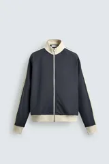 Sudadera de corte relajado en color azul marino con cuello alto y cierre frontal de cremallera. Presenta bandas laterales en color crema a contraste que recorren las mangas, puños y bajo en tejido rib, y bolsillos laterales de vivo.