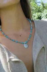 Gargantilla de cuentas de cristal en tonos tornasolados, violetas y oscuros, con dije central ovalado de metal plateado con diseño de escudo.
