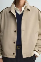 Cazadora color beige, estilo cropped, con cuello de solapa, manga larga con puños ajustables y trabillas en los hombros. Presenta bolsillos de vivo en la cadera y cierre frontal de botonadura.