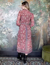 Vestido camisero midi de gasa estampada con flores rojas y blancas sobre fondo marrón. Tiene cuello con solapa, abertura central con botones, mangas largas y lazo de ajuste en la cintura.