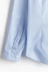 Camisa de tela azul claro con tratamiento de planchado fácil, cuello inglés, tapeta clásica y canesú en la espalda. Mangas largas con tapeta y botón, y cierre ajustable de botón en los puños. Bajo levemente redondeado. Corte ajustado que realza el contorno del cuerpo.
