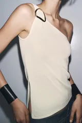 Top blanco de punto con escote asimétrico y aplique metálico dorado en el hombro.