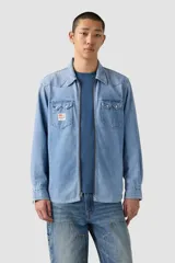 Camisa de jean azul claro, estilo western, con cierre frontal de cremallera, cuello clásico, dos bolsillos con solapa en el pecho y detalles de costuras en forma de "V" en el canesú. Incluye un parche decorativo en el bolsillo izquierdo.