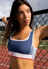 Top deportivo azul con contorno y tirantes blancos, banda inferior blanca con logo en rojo y borde inferior rojo.