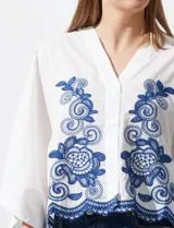 Blusa blanca con bordado floral azul, escote en V, manga japonesa y cierre frontal con botones.