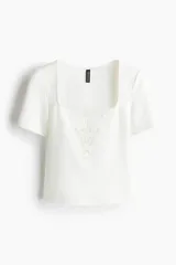 Top de punto color blanco con escote cuadrado y mangas cortas. Presenta un detalle decorativo en forma de V en broderie en el centro del busto.
