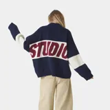 Sweater de punto grueso color azul marino con franja horizontal en el pecho que contiene la palabra "EME" en color bordó, bordeada en blanco. Las mangas son de color azul marino con una franja ancha color crema/blanco roto en la parte superior del brazo.