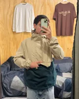 Campera rompeviento de diseño bicolor, con parte superior en tono arena y parte inferior en verde oscuro. Cuenta con capucha, cuello alto, cierre frontal parcial oculto y un amplio bolsillo frontal tipo canguro.