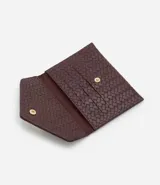 Billetera pequeña tipo sobre con textura trenzada tressê, confeccionada en cuero sintético. Presenta un cierre con botón a presión y un aplique metálico triangular decorativo en la solapa.
