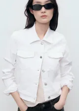 Chaqueta crop estilo denim de color blanco, confeccionada en algodón con elasticidad. Cuenta con cuello camisero, manga larga con puños abotonados, cierre frontal con botones y cuatro bolsillos (dos laterales y dos de parche con solapa en el pecho).