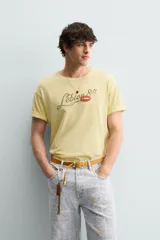Camiseta de corte relajado color amarillo con efecto lavado. Presenta cuello redondo, mangas cortas con dobladillo y un estampado frontal con texto y un parche decorativo.