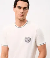 Remera de manga corta y cuello redondo color blanco crudo, con detalles de ribetes oscuros en cuello, mangas y ruedo, y un pequeño escudo bordado en el pecho.