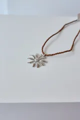 Collar con tiento trenzado color crema y dije de acero quirúrgico hipoalergénico con forma de sol o flor estilizada.