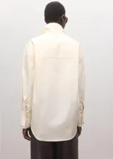 Blusa oversize de algodón en color marfil, con cuello camisero alto, manga larga con puños abotonados y cierre frontal de botonadura oculta.