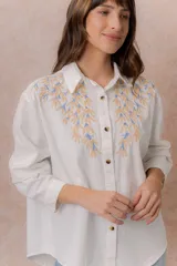 Camisa de denim azul con bordados florales en tonos amarillos y celestes sobre los hombros y el pecho. Presenta cuello clásico, cierre frontal con botones y mangas largas con puños.