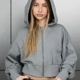 Campera con capucha color gris, de corte corto (cropped), con cierre frontal completo y mangas largas holgadas con puños elásticos.