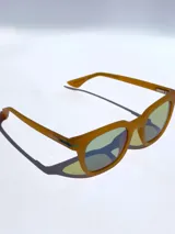 Lentes de sol de marco grueso color mostaza con acabado mate y cristales polarizados con reflejo verdoso. Presentan el logo de la marca grabado en la patilla.