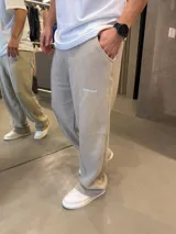 Pantalón deportivo de corte baggy y estilo oversize, color beige. Presenta costuras verticales decorativas en el frente, bolsillos laterales y un pequeño logo bordado en la pierna derecha.