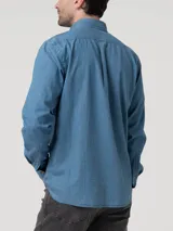 Camisa de manga larga color azul claro, confeccionada en tela tipo denim o chambray, con cuello abotonado (button-down) y un pequeño logo bordado de un pato en el pecho.