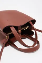 Bolso bandolera de tamaño pequeño de cuero marrón, con asas de mano, asa ajustable y logo grabado en dorado en el frente.