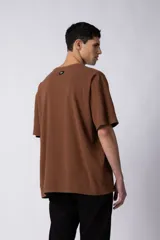 Remera de corte oversized en color marrón, confeccionada en tejido de algodón con textura de micropanal. Presenta cuello redondo con detalle de costura en forma de X y mangas cortas de estilo caído.