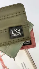 Billetera corta de diseño capitoneado con cierre perimetral y solapa frontal. Cuenta con compartimentos internos para tarjetas y billetes, e incluye una correa de mano desmontable.