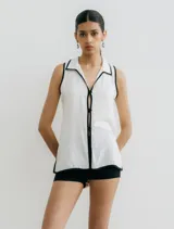 Blusa blanca sin mangas con cuello camisero y ribete negro en cuello, sisas y ruedo. Presenta abertura en los costados y cierre frontal con botones negros.
