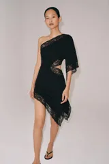 Vestido negro de escote asimétrico con un solo hombro y manga larga ancha. Presenta detalles de encaje negro en el escote, el ruedo asimétrico y un corte 'cut out' en el lateral del torso.