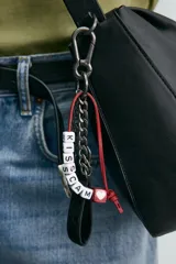 Bolso tipo bandolera de diseño minimalista en color negro, con cierre de cremallera superior y asa de hombro ajustable. Incluye un detalle decorativo de charms metálicos en uno de los laterales.