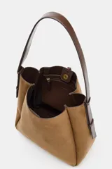 Bolso tipo saca de gran tamaño confeccionado en material efecto gamuza color verde oliva. Presenta un diseño estructurado con pespuntes decorativos, una anilla metálica dorada en el frente y cierre mediante botón imantado. Incluye un asa de hombro ajustable y bolsillo interior con cremallera.