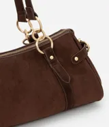 Cartera mediana de diseño alargado y estructura firme, confeccionada en gamuza sintética de color marrón. Cuenta con doble asa fina sujeta por argollas metálicas doradas, cierre superior y detalles de solapas decorativas con tachas en los laterales.