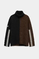 Polera de cuello alto confeccionada en lana lambswool, con diseño bicolor en negro y marrón. Presenta mangas largas con franjas verticales en gris y un parche circular bordado en la manga derecha.