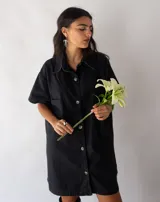Vestido camisero corto negro de denim, con cuello clásico, cierre frontal con botones, mangas cortas, bolsillos cuadrados en el pecho y corte recto.