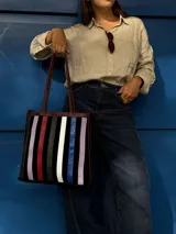 Cartera tipo tote de cuero con diseño de franjas verticales multicolores en tonos tierra, rojo y azul. Cuenta con correas trenzadas de cuero y cierre superior.