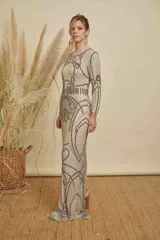 Vestido largo de fiesta color beige, con mangas largas y cuello redondo. Presenta un intrincado diseño bordado con canutillos y lentejuelas en tonos plata y gris, creando patrones geométricos y ornamentales en toda la prenda.
