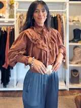 Blusa de color marrón con diseño romántico, cuello alto con lazo ajustable y volados tipo shabot en el frente. Presenta mangas largas abullonadas con puños ajustados y una textura suave y liviana.