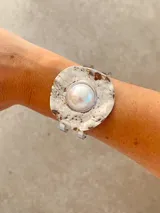Brazalete rígido de diseño circular con acabado plateado texturizado e irregular, destacando una perla central grande. Presenta un estilo moderno y llamativo.