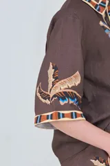 Camisa de lino marrón con estampado de hojas de palmera y detalles geométricos en color azul, celeste y naranja en el cuello, la tapeta y los puños. Tiene mangas cortas y corte recto.