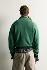 Sudadera regular fit color verde con cuello subido y cierre frontal de cremallera corta en tono naranja a contraste. Cuenta con mangas largas raglán, bolsillos frontales tipo canguro y puños y cintura en canalé de color azul marino.