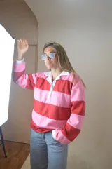 Sweater estilo rugby a rayas horizontales rosas y rojas con cuello blanco y cierre parcial con botones.