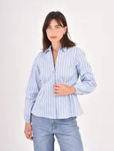 Camisa de corte entallado con pinzas en la cintura, cuello clásico y mangas largas con puños elásticos. Presenta un diseño a rayas verticales finas en color celeste sobre fondo claro.