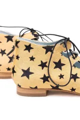 Semibotín de piel de pony con estampado de estrellas negras sobre fondo ocre. Presenta un diseño con abertura escotada, cierre de cordones negros a contraste y punta afilada. Incluye cuña interna y suela de cuero.