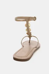 Sandalia plana color beige con tira para el dedo y tira delantera con detalle de abalorios metálicos. Cierre mediante tira con hebilla en el tobillo y acabado en punta redonda.