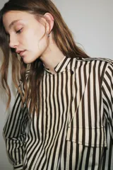 Camisa tipo bomber confeccionada en algodón, con diseño de rayas verticales en tonos marrón y blanco. Presenta cuello perkins, manga larga con puños, bolsillo de plastrón con solapa en el frente, bajo elástico y cierre frontal con botones.
