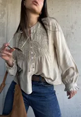 Camisa beige de manga larga con detalles bordados en el frente, cuello redondo y cierre frontal con botones. Las mangas presentan puños con volados.