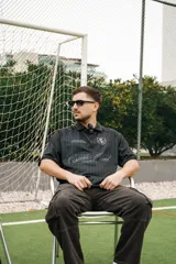 Camiseta de fútbol estilo boxy de manga corta y cuello polo, en color negro con sutiles rayas verticales. Presenta inscripciones estampadas y escudo bordado en el pecho.