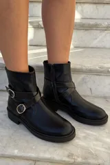Bota corta de cuero negro con acabado brillante, diseño de caña media con cierre lateral y tiras decorativas con hebillas metálicas plateadas.