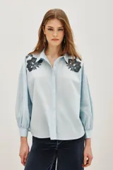 Camisa blanca de algodón con cuello camisero y mangas largas abullonadas con puños ajustados. Presenta detalles bordados en los hombros con motivos vegetales y aplicaciones brillantes.