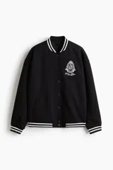 Campera de béisbol negra de efecto neopreno, con hombros caídos, cuello, puños y bajo acanalados con rayas blancas. Presenta cierre frontal con botones a presión, bolsillos ribeteados y un escudo bordado en el pecho.